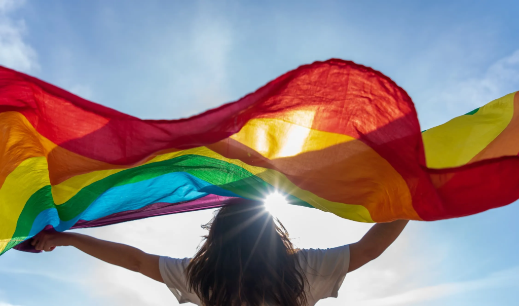 Woman stands with her back to the camera, a rainbow pride flag held between her hands and raised over her hear. It is a sunny day.